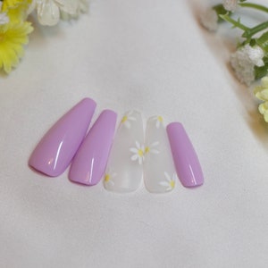 May include: Five lavender-colored press-on nails with a white daisy design. The nails are arranged in a row on a white surface.