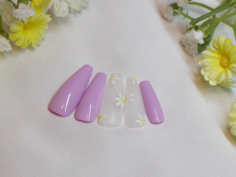 May include: Five lavender-colored press-on nails with a white daisy design. The nails are arranged in a row on a white surface.