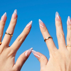 Puede incluir: Dos manos con u&ntilde;as largas y almendradas pintadas con un dise&ntilde;o de espiral blanco sobre una base nude. Las manos llevan anillos de oro.