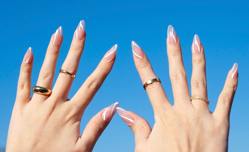 Puede incluir: Dos manos con u&ntilde;as largas y almendradas pintadas con un dise&ntilde;o de espiral blanco sobre una base nude. Las manos llevan anillos de oro.