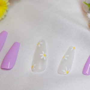 May include: Set of six false nails, five with a white base and small yellow daisy designs, and one solid light purple nail.