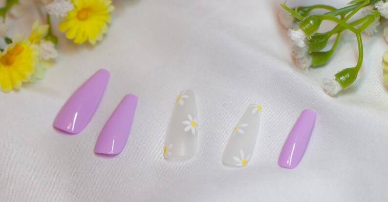 May include: Set of six false nails, five with a white base and small yellow daisy designs, and one solid light purple nail.