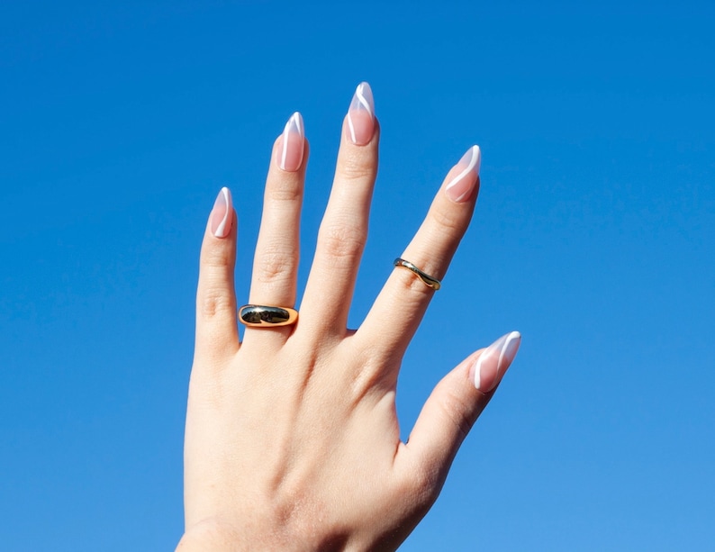 Puede incluir: Una mano con u&ntilde;as largas y almendradas pintadas con un dise&ntilde;o de punta blanca. Las u&ntilde;as est&aacute;n pintadas de un color rosa claro. La mano lleva dos anillos de oro.
