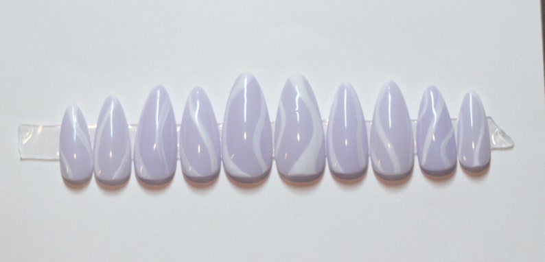 Puede incluir: Conjunto de diez u&ntilde;as postizas de color lavanda con dise&ntilde;os de remolinos blancos.