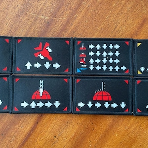 May include: Eight black rectangular patches with various red and white graphics. Designs include arrows, a jet, a mushroom cloud, and a structure with three smokestacks. Each patch has a white arrow pattern.