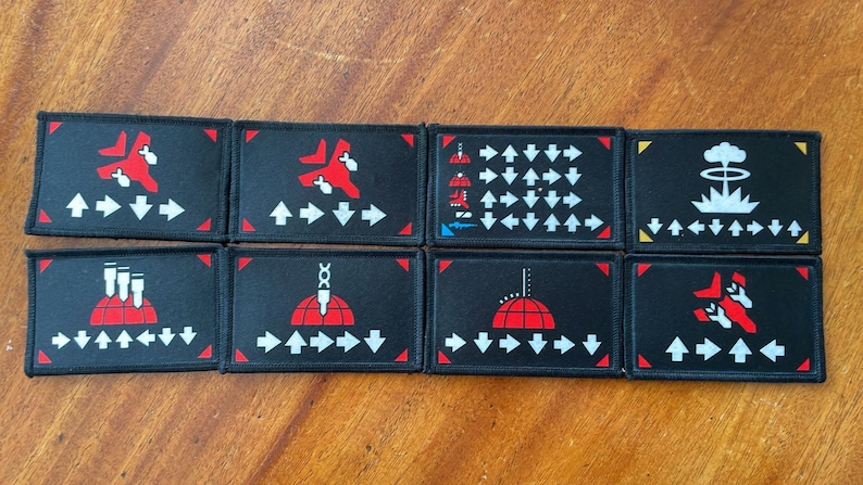 May include: Eight black rectangular patches with various red and white graphics. Designs include arrows, a jet, a mushroom cloud, and a structure with three smokestacks. Each patch has a white arrow pattern.
