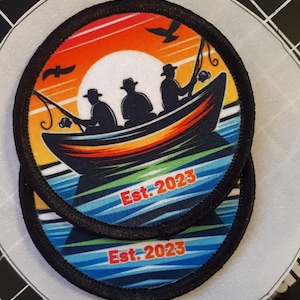 May include: Two circular patches with a fishing theme. The patches feature a boat with three silhouetted figures fishing against a sunset backdrop. The text "Est. 2023" is printed in red. The patches have a black border.