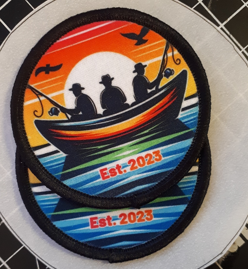 May include: Two circular patches with a fishing theme. The patches feature a boat with three silhouetted figures fishing against a sunset backdrop. The text "Est. 2023" is printed in red. The patches have a black border.