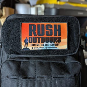 May include: A black backpack with a rectangular patch that reads "RUSH OUTDOORS" in bold, black letters over an orange and yellow gradient. The patch also includes the text "JOIN ME ON THE JOURNEY" and social media handles.