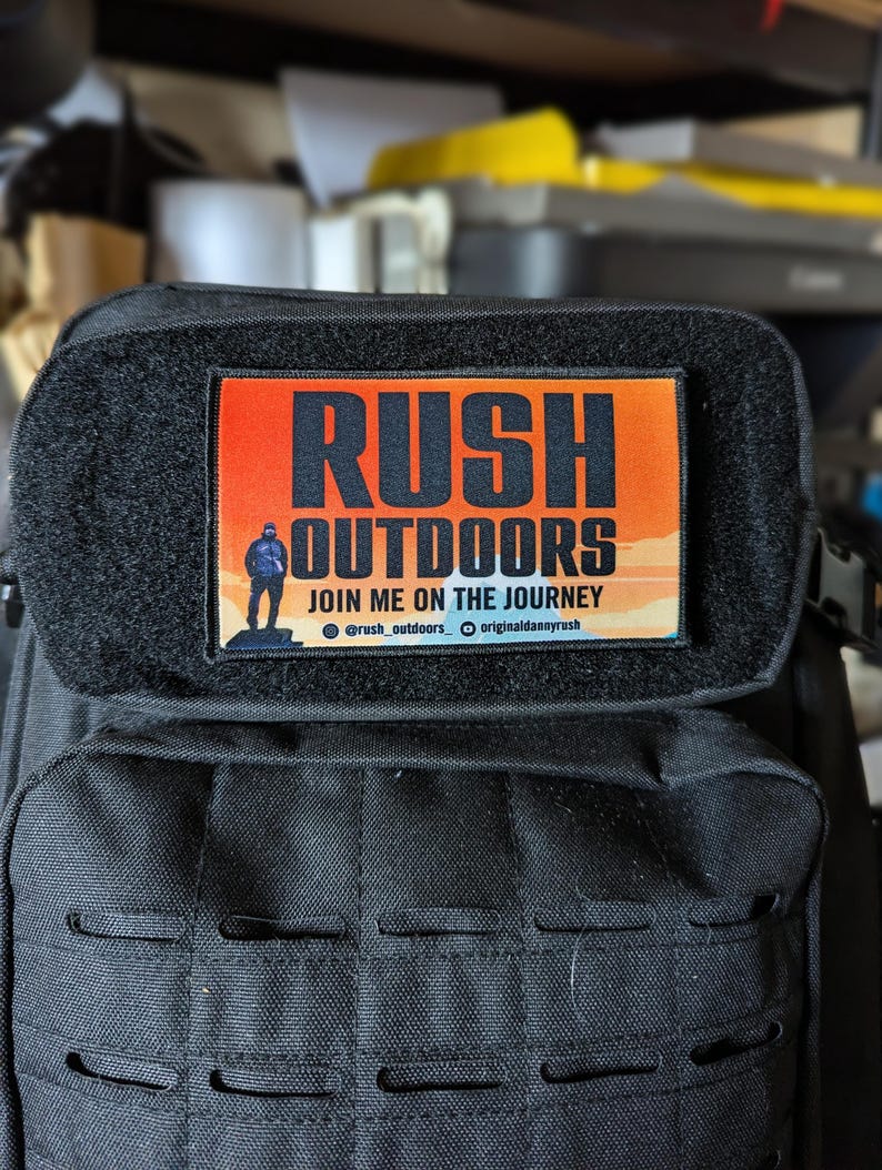 May include: A black backpack with a rectangular patch that reads "RUSH OUTDOORS" in bold, black letters over an orange and yellow gradient. The patch also includes the text "JOIN ME ON THE JOURNEY" and social media handles.