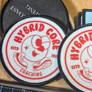 May include: A collection of round embroidered patches. The primary patch features a red and white design with the words "HYBRID CORE COACHING" and a cartoon character. The patches have black borders and are on a tan surface.
