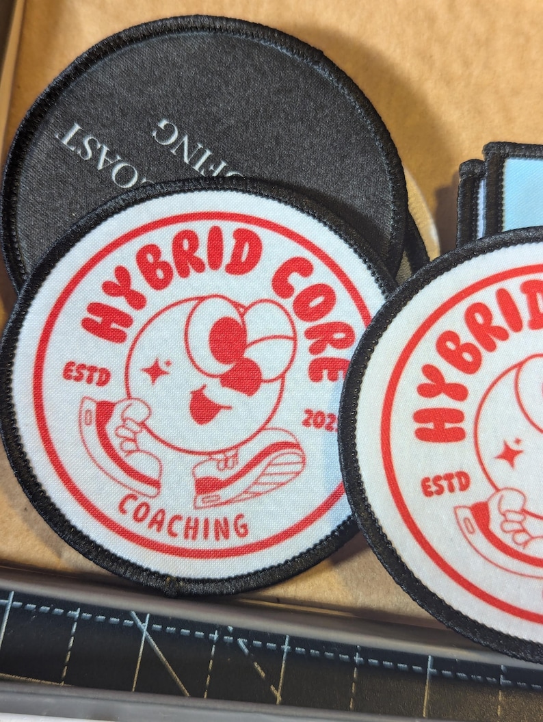 May include: A collection of round embroidered patches. The primary patch features a red and white design with the words "HYBRID CORE COACHING" and a cartoon character. The patches have black borders and are on a tan surface.
