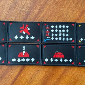 May include: Eight black rectangular patches with various red and white graphic designs. Each patch has a different design, including arrows, a mushroom cloud, and other abstract shapes. The patches are arranged in two rows of four.