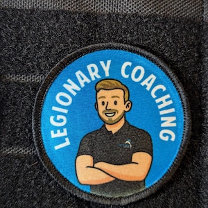 May include: A round patch with the words "LEGIONARY COACHING" in white text on a blue background. The patch features a cartoon illustration of a man with crossed arms, wearing a black shirt. The patch is set against a dark gray, textured background.
