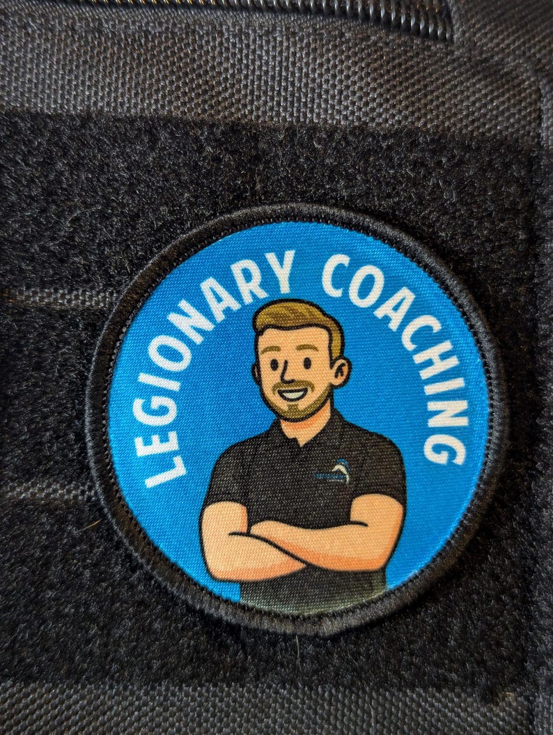 May include: A round patch with the words "LEGIONARY COACHING" in white text on a blue background. The patch features a cartoon illustration of a man with crossed arms, wearing a black shirt. The patch is set against a dark gray, textured background.