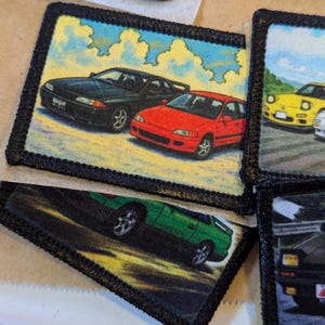 May include: Embroidered patches featuring colorful illustrations of classic cars. The patches have a black border and depict various vehicles against a backdrop of sky and clouds. The cars are in shades of black, red, green, and yellow.