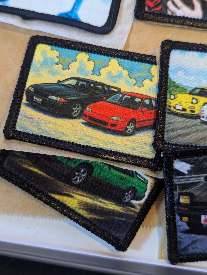 May include: Embroidered patches featuring colorful illustrations of classic cars. The patches have a black border and depict various vehicles against a backdrop of sky and clouds. The cars are in shades of black, red, green, and yellow.