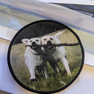 May include: Round patch featuring two white dogs joyfully carrying a stick in their mouths. The dogs are set against a backdrop of green grass and a soft, blurred background. The patch has a black border.