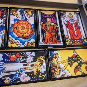 May include: A collection of tarot card patches featuring vibrant illustrations. The cards depict figures in regal attire, celestial symbols, and scenes of fortune. Each patch has a black border, and the cards are arranged in a tray.