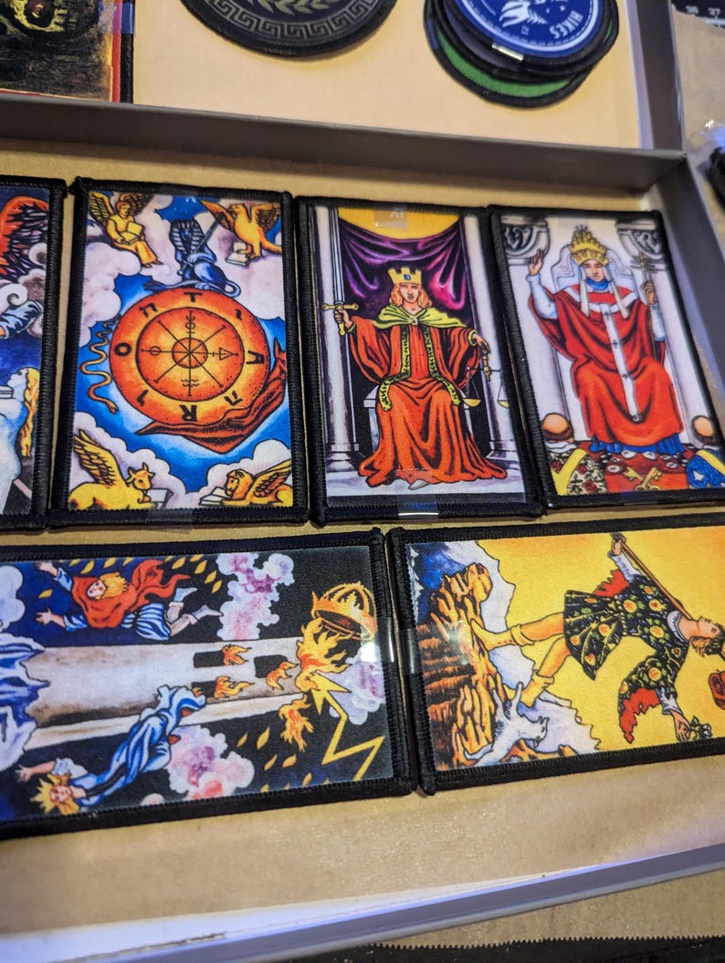 May include: A collection of tarot card patches featuring vibrant illustrations. The cards depict figures in regal attire, celestial symbols, and scenes of fortune. Each patch has a black border, and the cards are arranged in a tray.