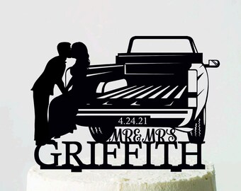 May include: A black cake topper with a silhouette of a couple kissing in front of a truck. The date 4.24.21 and the names Mr & Mrs Griffith are displayed below the truck. The white cake sits on a white cake stand.