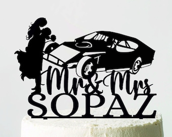 Könnte beinhalten: Ein schwarzer Tortenaufsatz mit einer Silhouette eines Paares, das sich neben einem klassischen Auto umarmt. Die Worte "Mrs & Mrs SOPAZ" sind unter dem Auto zu sehen. Der Aufsatz befindet sich auf einem weiß glasierten Kuchen.