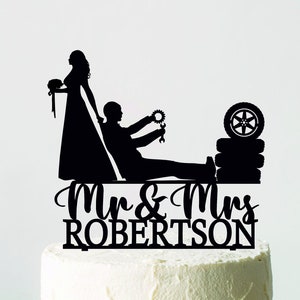 May include: A black cake topper featuring a bride and groom silhouette, with the groom holding tools and a tire. The topper is placed on a white cake with the words "Mr & Mrs ROBERTSON" in script.