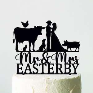 Pode incluir: Um topo de bolo preto com a silhueta de uma noiva e um noivo abraçados, com animais de fazenda, incluindo uma vaca, um porco, um cão, um gato e uma galinha. As palavras "Mr & Mrs Easterby" estão abaixo. O topo está em cima de um bolo branco gelado.