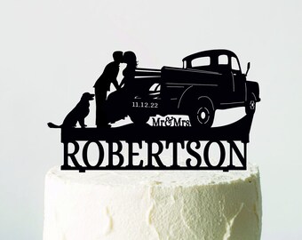 May include: A black cake topper depicting a couple kissing beside a vintage truck, with a dog. The topper includes the date 11.12.22, and the name "ROBERTSON" below. The cake is on a white cake stand.