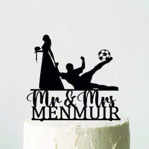 May include: A wedding cake with white frosting, presented on a white stand. The cake is decorated with a black silhouette cake topper depicting a bride, groom, and a football. The topper reads "Mr & Mrs MENMUIR".