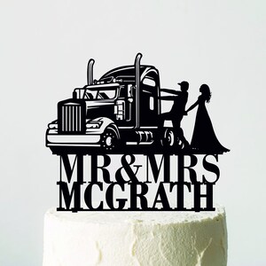 May include: Black laser-cut cake topper featuring a semi-truck, a bride, and a groom. The topper reads "MR & MRS MCGRATH" in bold, capital letters. The cake is white and sits on a white cake stand.