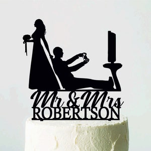 May include: A black silhouette cake topper featuring a bride and groom. The groom is depicted playing a video game, while the bride stands beside him. The topper includes the text "Mr & Mrs ROBERTSON" in a script font. The cake is white and sits on a white cake stand.