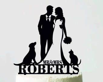 May include: A wedding cake with a white frosting, topped with a black silhouette cake topper. The topper features a bride and groom, a dog, a cat, and the text "MR & MRS ROBERTS". The cake sits on a white cake stand.