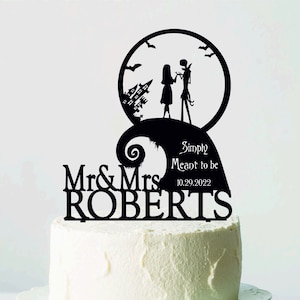 Pu&ograve; includere: Un cake topper nero con la silhouette di una coppia, una citt&agrave; spettrale e pipistrelli, con le parole "Simply Meant to be" e la data 10.29.2022. Il topper &egrave; su una torta glassata bianca su un alzatina bianca.