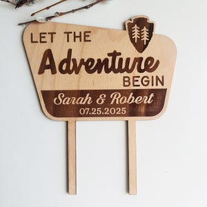 May include: Wooden wedding cake topper with the words "LET THE ADVENTURE BEGIN" and the names "Sarah & Robert" along with the date 07.25.2025. The topper has a shield with two trees and two wooden stakes for insertion into the cake.