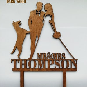 May include: A dark wood wedding cake topper featuring a silhouette of a couple, a dog, and the text "MR & MRS THOMPSON". The topper is made of dark wood and is designed to be inserted into a cake.