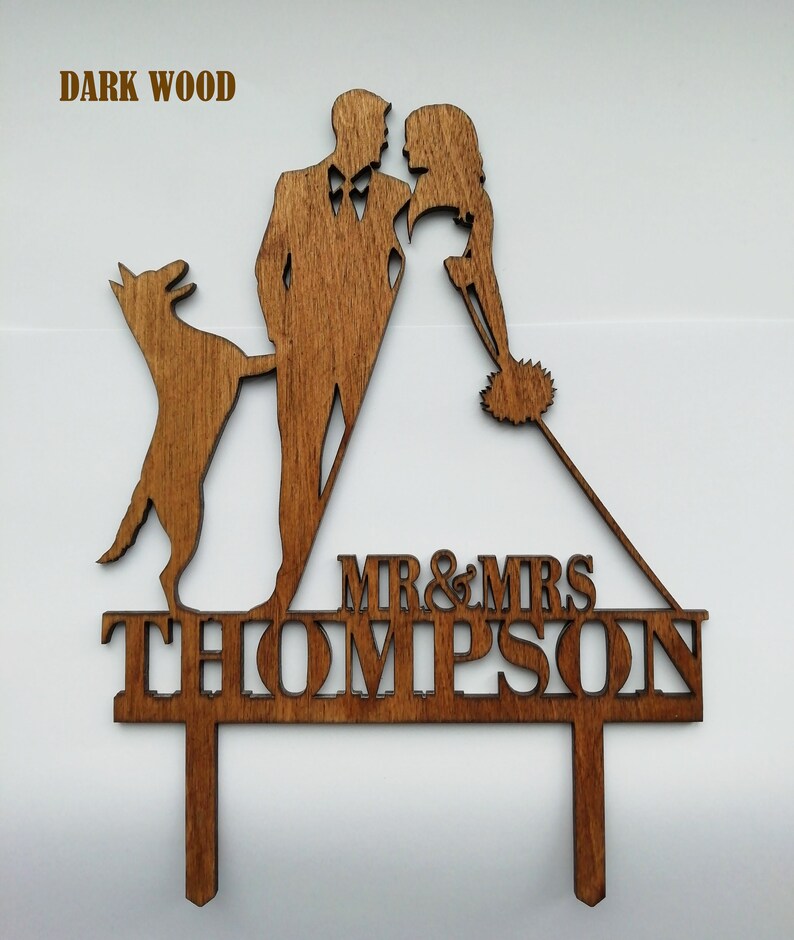 May include: A dark wood wedding cake topper featuring a silhouette of a couple, a dog, and the text "MR & MRS THOMPSON". The topper is made of dark wood and is designed to be inserted into a cake.