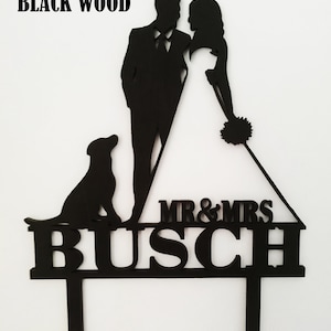 Pu&ograve; includere: Cake topper nuziale in legno nero con sagome di sposi e cane. Il topper include la scritta "MR & MRS" sopra il cognome "BUSCH". Le parole "BLACK WOOD" sono in alto.