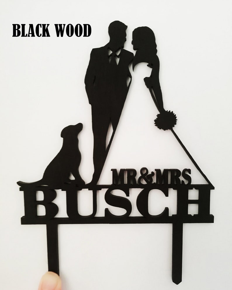 Pu&ograve; includere: Cake topper nuziale in legno nero con sagome di sposi e cane. Il topper include la scritta "MR & MRS" sopra il cognome "BUSCH". Le parole "BLACK WOOD" sono in alto.