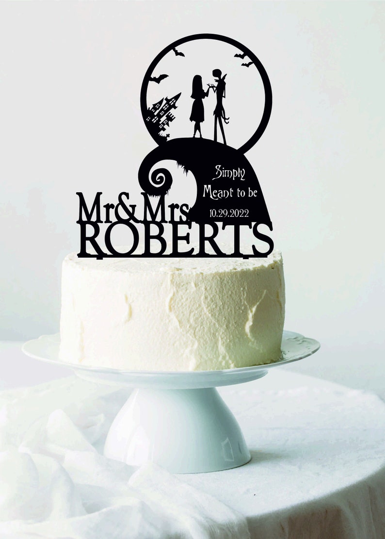 Pu&ograve; includere: Un cake topper nero con la silhouette di una coppia, una citt&agrave; spettrale e pipistrelli, con le parole "Simply Meant to be" e la data 10.29.2022. Il topper &egrave; su una torta glassata bianca su un alzatina bianca.