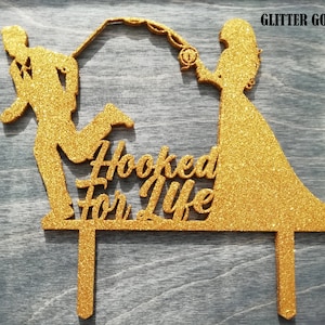 Pu&ograve; includere: Un cake topper dorato glitterato con la silhouette di una coppia, con l'uomo che corre e la donna che tiene una canna da pesca. Il testo "Hooked For Life" &egrave; al centro. Il testo "Glitter Gold" &egrave; nell'angolo in alto a destra.