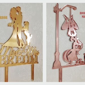 May include: Two mirrored acrylic cake toppers. One is gold and features a couple with a child and the text "Mr & Mrs BREHM". The other is rose gold and depicts a witch with a cat and a street lamp.