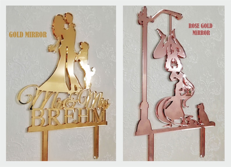 May include: Two mirrored acrylic cake toppers. One is gold and features a couple with a child and the text "Mr & Mrs BREHM". The other is rose gold and depicts a witch with a cat and a street lamp.