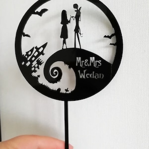 Pu&ograve; includere: Topper per torta nuziale in acrilico nero con sagome di una coppia, una casa stregata e pipistrelli. Il design include il testo "Mr & Mrs Wedan" all'interno di una cornice circolare, perfetto per un matrimonio a tema.