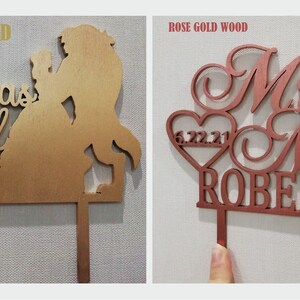 May include: Two wooden cake toppers. One is gold-colored with the words "Tale as Old as Time" and a silhouette of a couple. The other is rose gold with "Mr & Mrs ROBERTS" and a heart with the date 6.22.21.