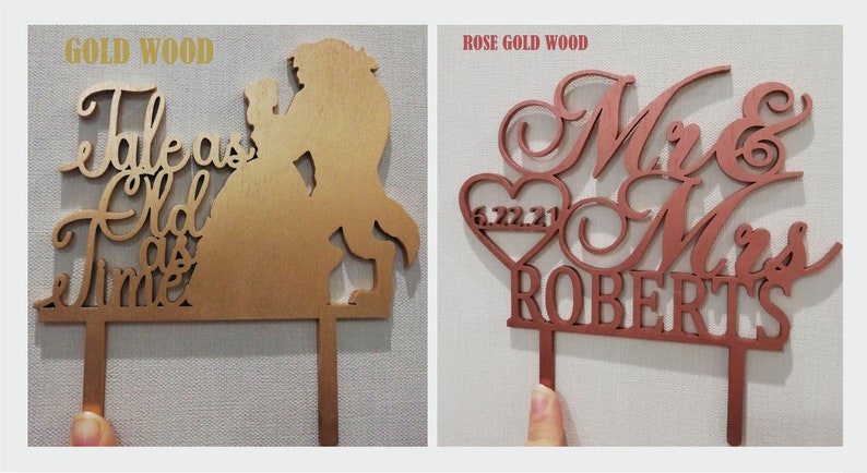 May include: Two wooden cake toppers. One is gold-colored with the words "Tale as Old as Time" and a silhouette of a couple. The other is rose gold with "Mr & Mrs ROBERTS" and a heart with the date 6.22.21.