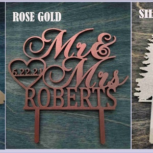 Pu&ograve; includere: Tre cake topper in legno tagliati al laser. Il topper dorato presenta una coppia e il testo "Once Upon a Dream". Il topper oro rosa dice "Mr & Mrs ROBERTS" con un cuore e una data. Il topper argentato mostra montagne, un albero e il testo "Our adventure continues."