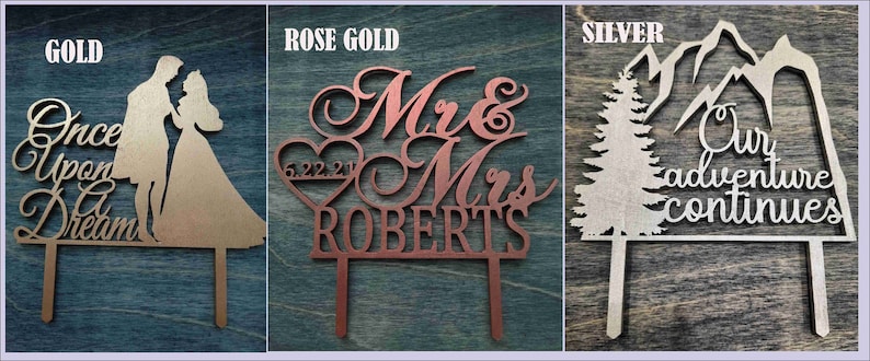 Pu&ograve; includere: Tre cake topper in legno tagliati al laser. Il topper dorato presenta una coppia e il testo "Once Upon a Dream". Il topper oro rosa dice "Mr & Mrs ROBERTS" con un cuore e una data. Il topper argentato mostra montagne, un albero e il testo "Our adventure continues."