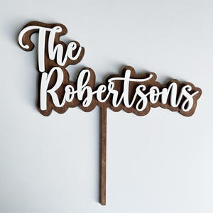 May include: A wooden cake topper with the words "The Robertsons" in a white, cursive font. The letters are layered on a dark brown wooden base with a scalloped edge. A thin wooden stick extends from the bottom for insertion into a cake.