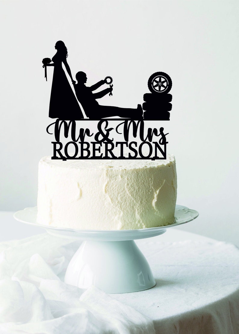 May include: A black cake topper featuring a bride and groom silhouette, with the groom holding tools and a tire. The topper is placed on a white cake with the words "Mr & Mrs ROBERTSON" in script.